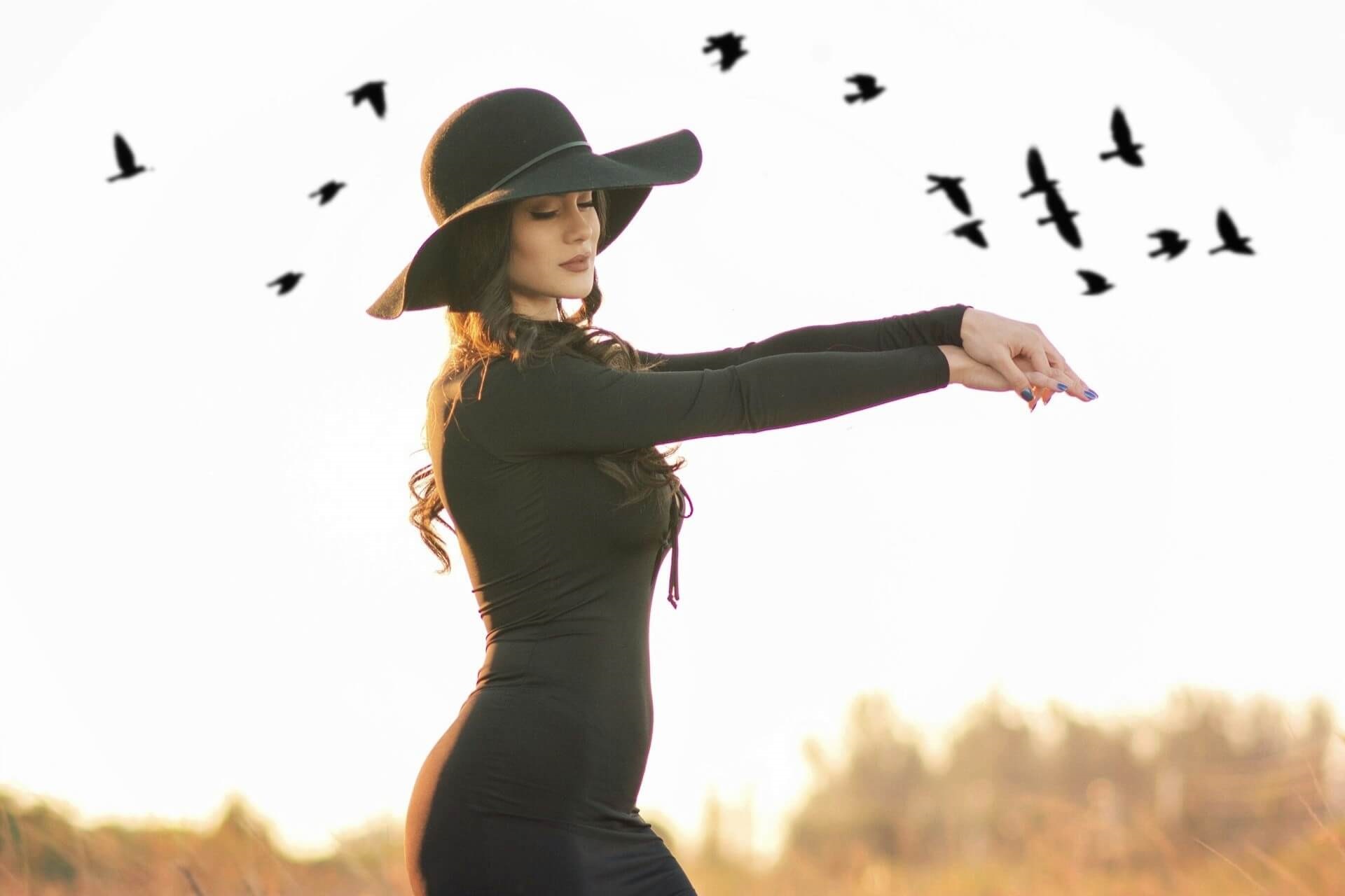 Kobieta w czarnym kapeluszu i sukience na tle nieba z lecącymi ptakami – wiosenne wróżby i ornitomancja
